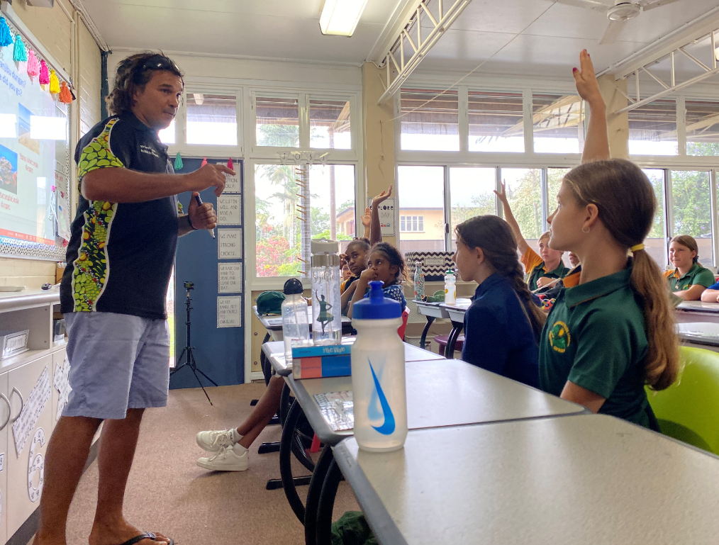 Juan Walker stands in front of a whiteboard at the front of a classroom facing students, some with hands in the air.